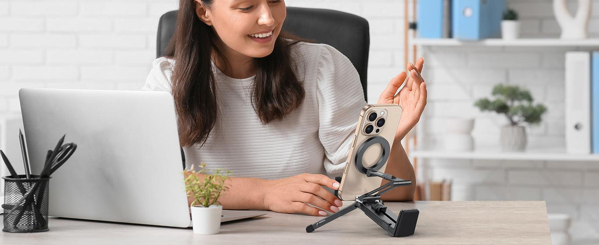Woman working at office desk with portable MagSafe magnetic phone tripod supporting her iPhone beside laptop.